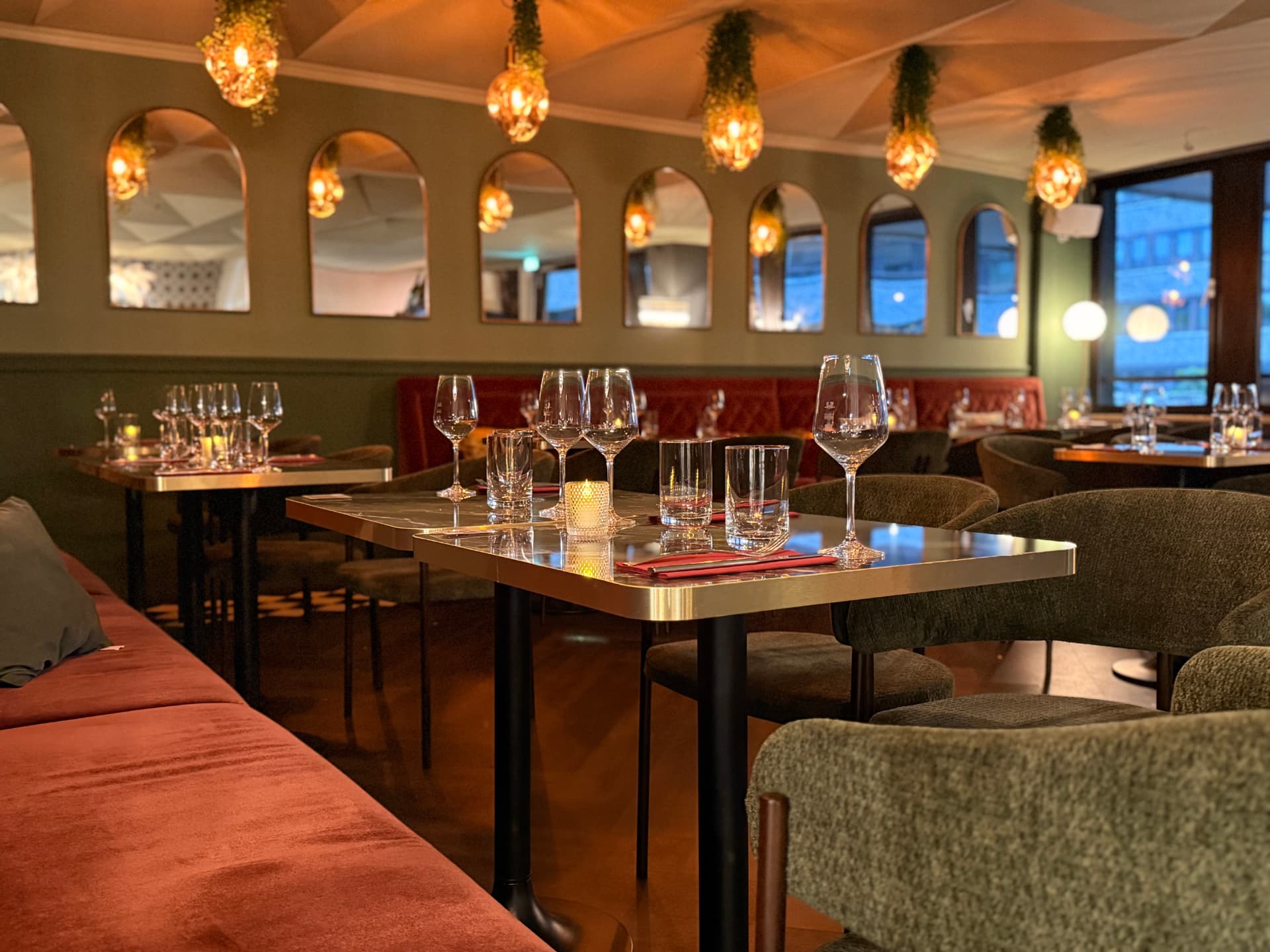 Gastraum des De Blanc am Abend: gedeckte Marmortische mit Weingläsern und Kerzenlicht, rostrote Polsterbank, olivgrüne Wand mit Rundbogenspiegeln und warme Hängeleuchten